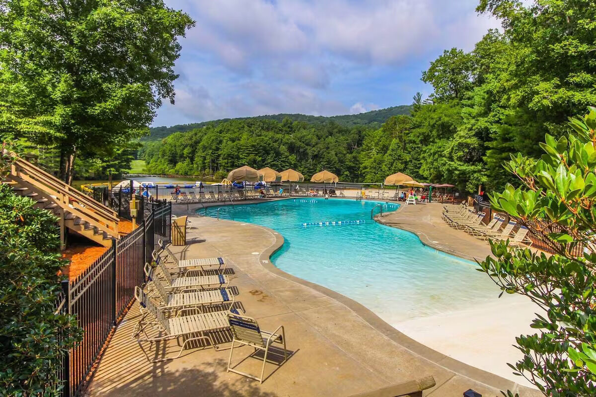 Community pool with a fresh deck, lounge chairs along the edge, sun umbrellas, and a wooded mountain backdrop